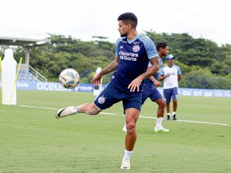 Jogador de futebol treina controlando a bola no pé em um campo de treino. Ele está usando o uniforme completo do Bahia, azul e branco, e está focado no movimento da bola para a Copa do Nordeste