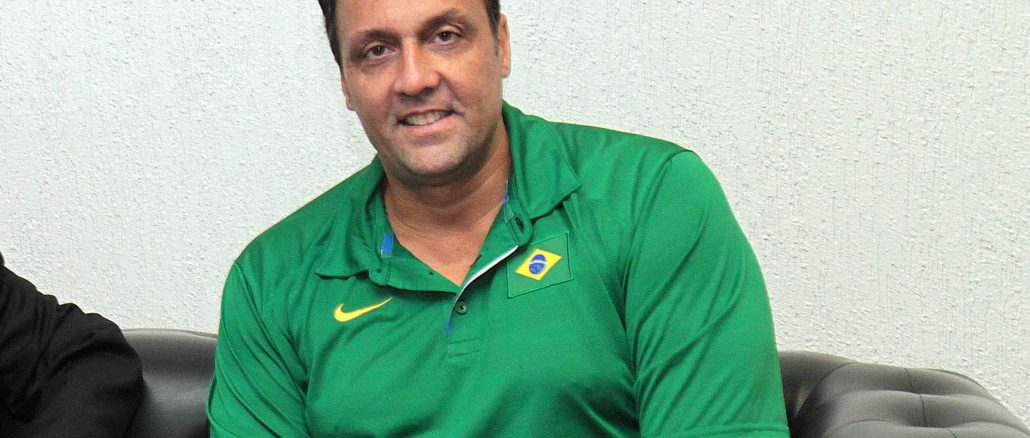 Pampa, ex-jogador de vôlei, vestido com polo verde da seleção brasileira, sentado e sorrindo.